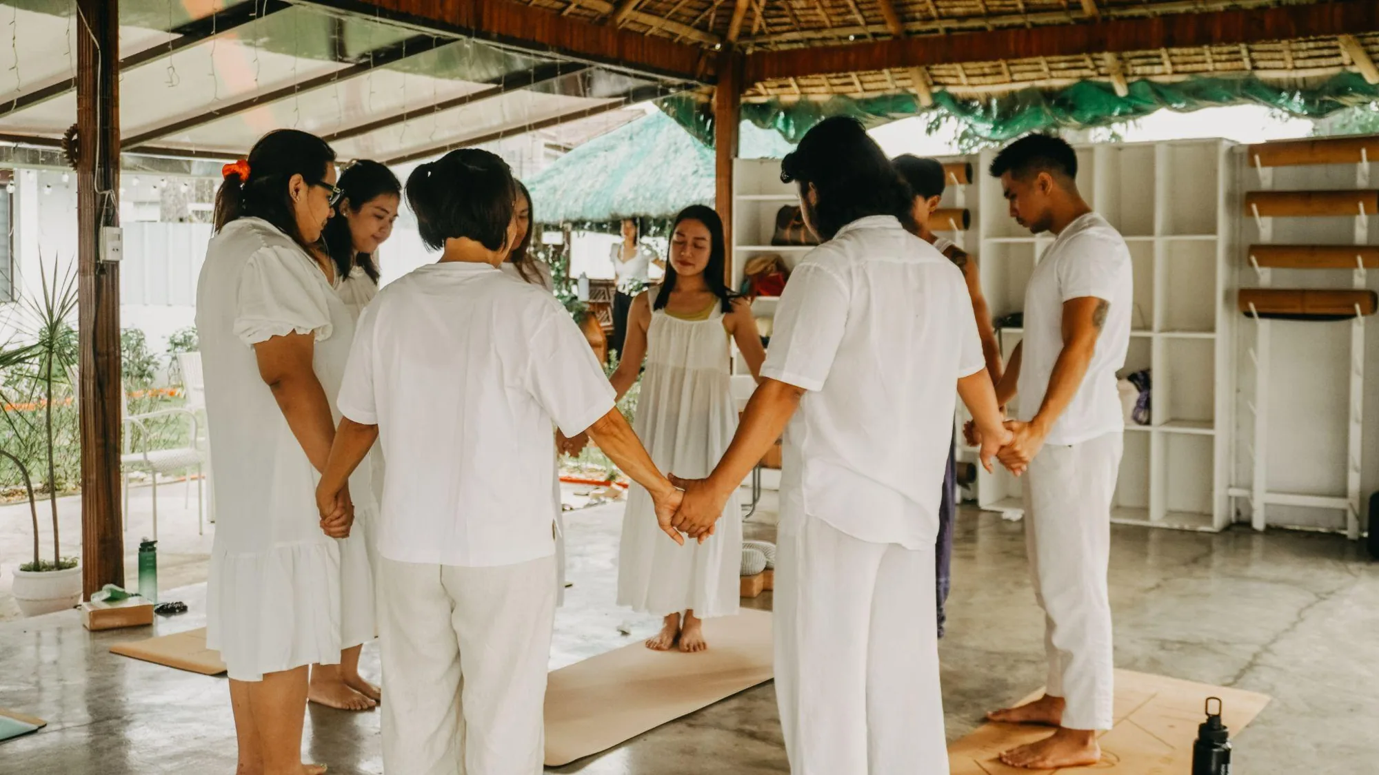 A Tallaro group in white standing in a circle, holding hands on yoga mats in the open pavilion
