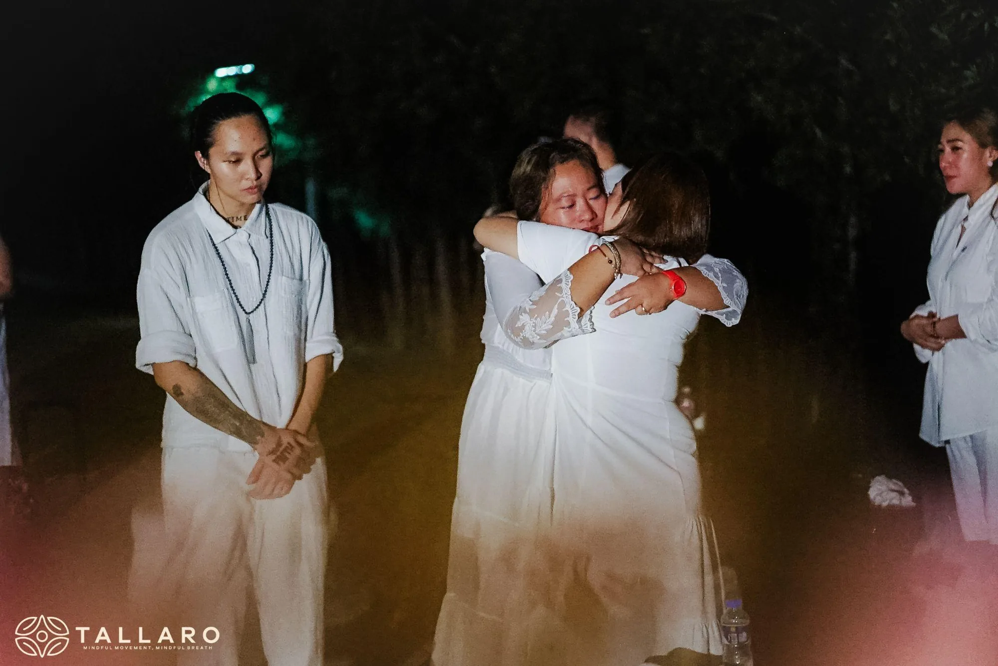 Two participants sharing a long embrace at night while others in white stand quietly beside them