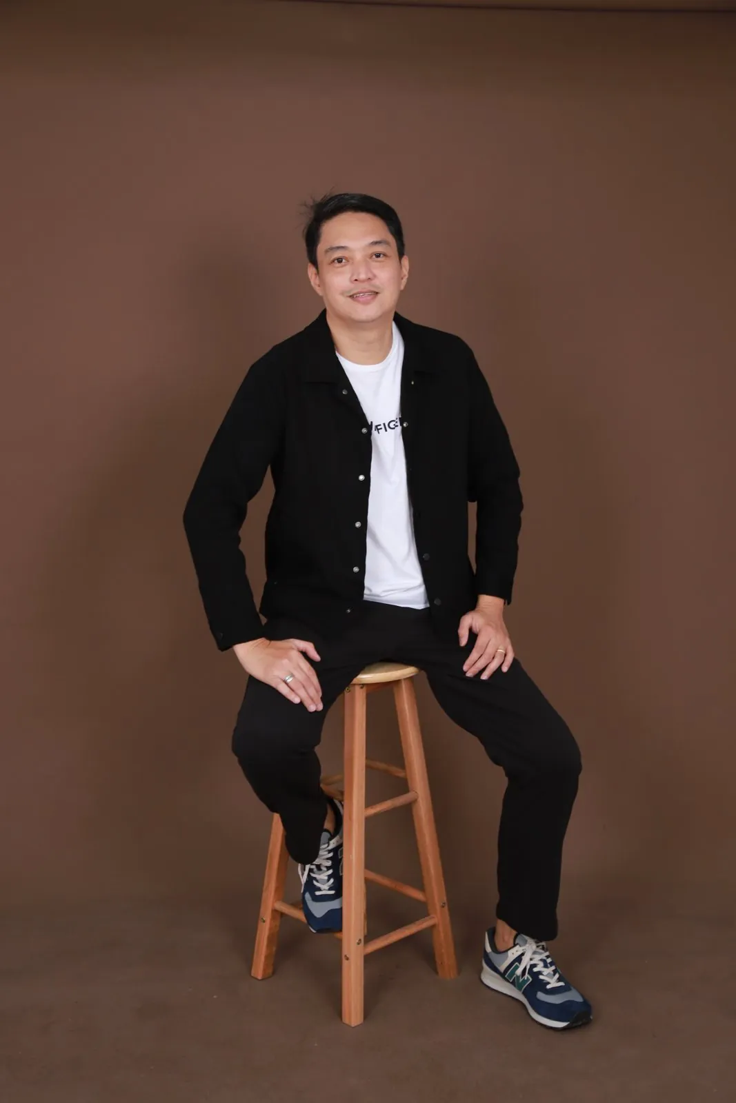 Clarence Gil seated on a wooden stool in a black jacket against a brown studio backdrop