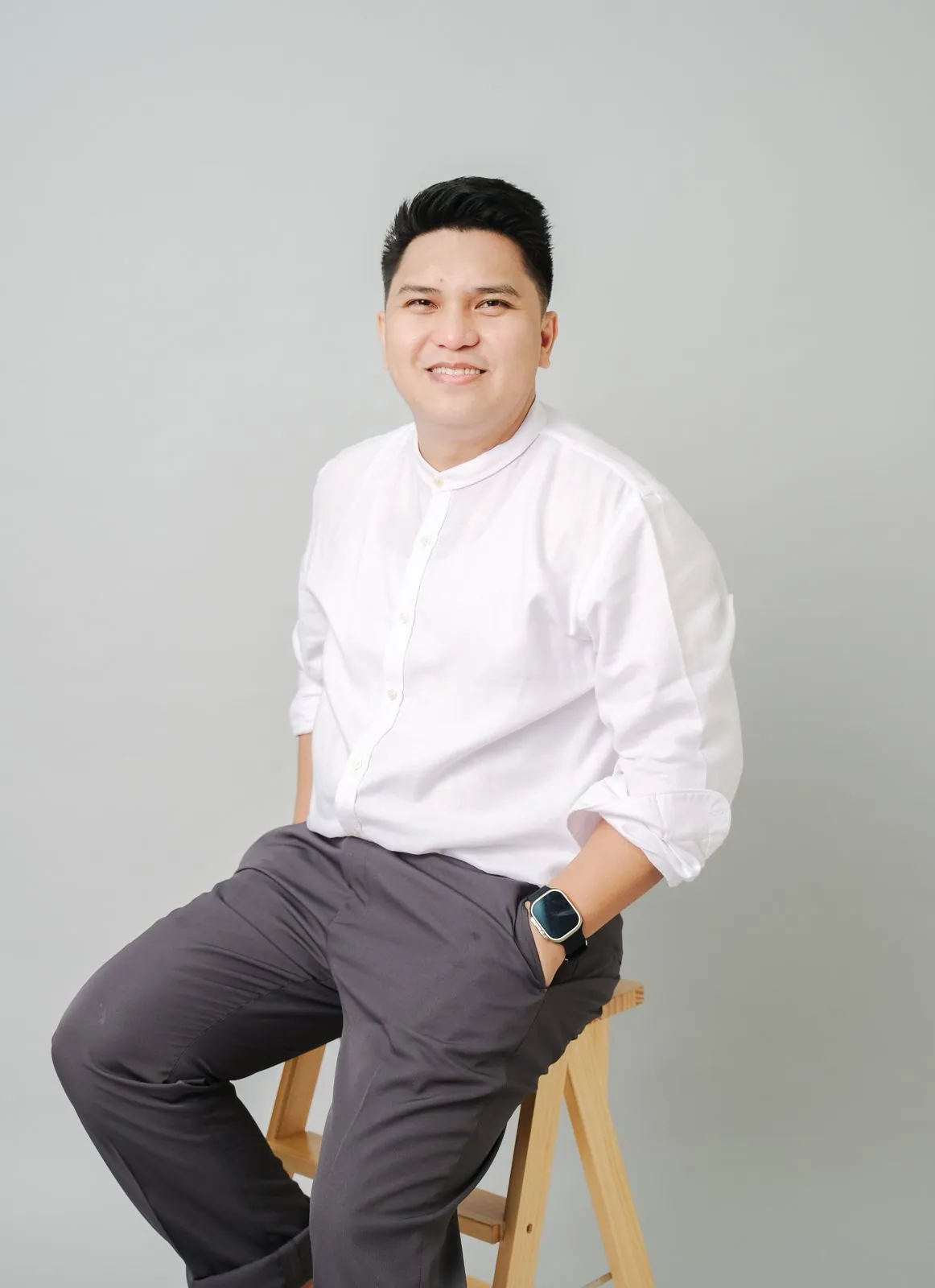Mau Magallanes seated on a wooden stool in a white shirt against a grey studio backdrop